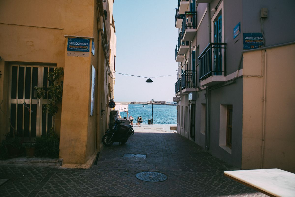 A narrow stone-paved lane in Chania old town opening onto the harbour, with Greek street signs on the walls