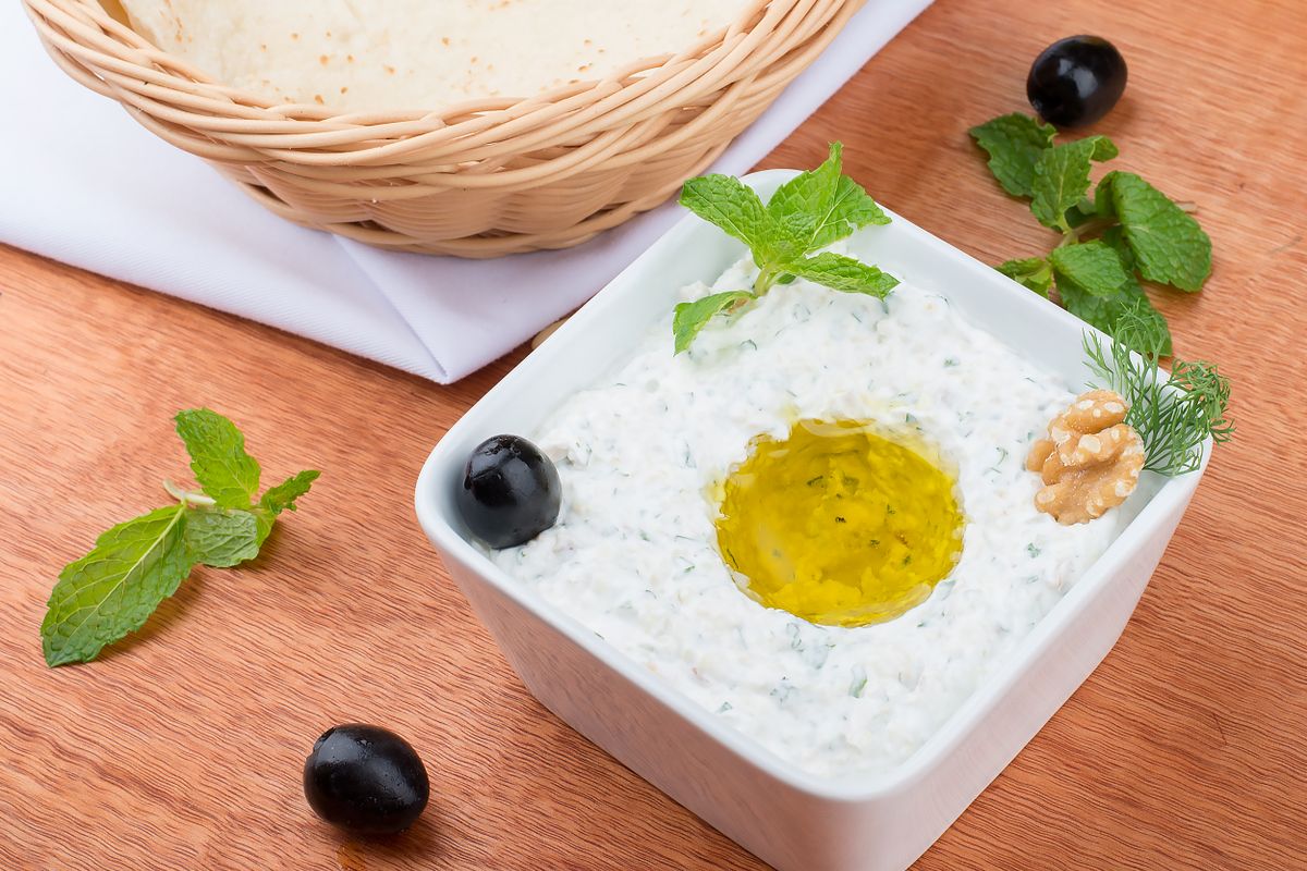 A bowl of tzatziki topped with olive oil and mint beside a basket of flatbread and black olives on a wooden taverna table