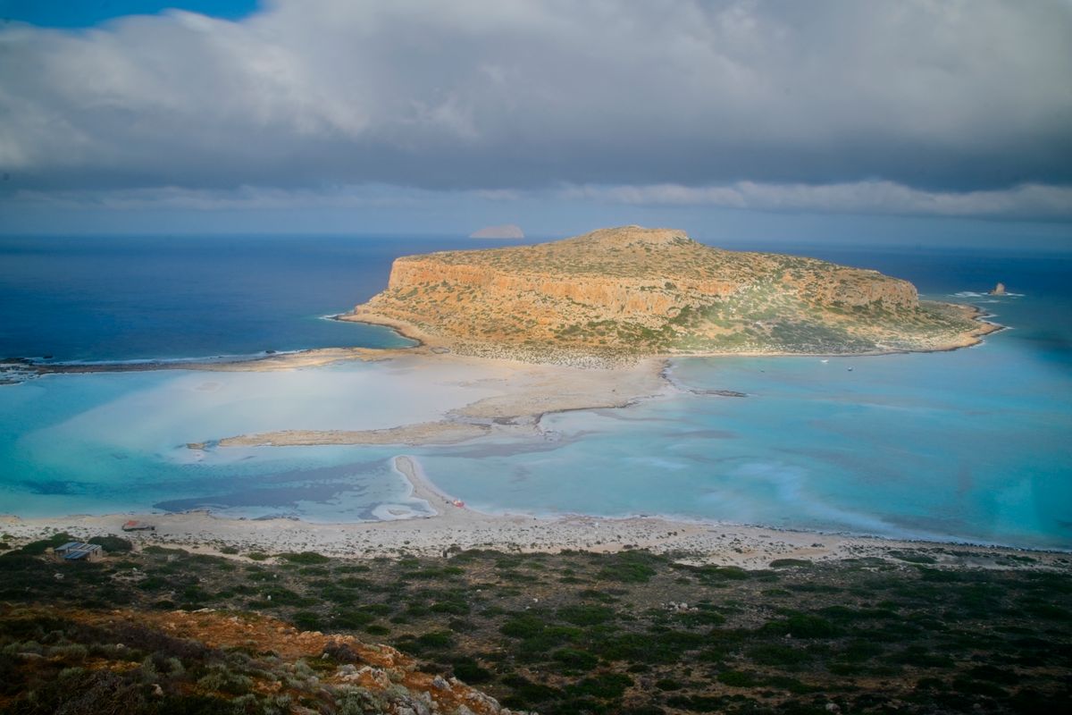 The Balos lagoon seen from above, a white sandbar curving toward a rocky headland with turquoise shallows on either side
